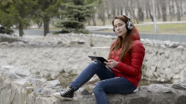 En tonårs flicka med hörlurar och en tablett i hennes händer vilar och lyssnar på musik sitter nära en tom fontän i staden våren Park i en röd jacka. Porträtt. 29,97 fps. — Stockvideo