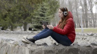 Kulaklıkta güzel bir genç kız müzik dinler ve şarkı söylüyor, kırmızı bir ceket şehir bahar Park taşlar üzerinde oturan ve kafasına kırmızı saç ile. Portre. Kapat şunu. 29,97 kare/sn fps.