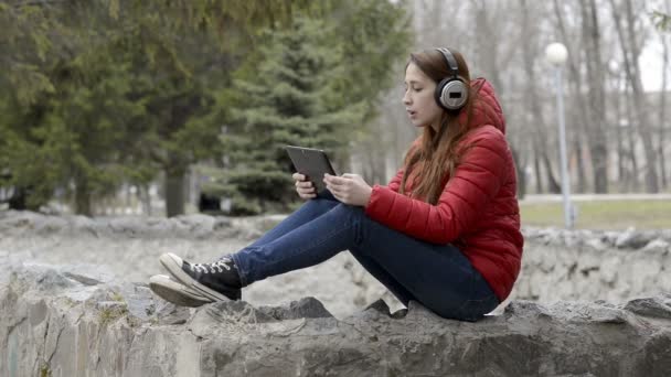 Una bella ragazza in cuffia ascolta musica e canta, seduta su pietre nel parco primaverile della città in giacca rossa e con i capelli rossi in testa. Ritratto. Chiudete. 29,97 fps . — Video Stock