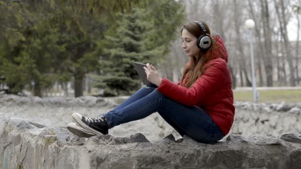 Una chica con grandes auriculares en la cabeza y una tableta en las manos sentada en las rocas en el parque de primavera de la ciudad con una chaqueta roja, escuchando música y bailando. Retrato. De cerca. 29,97 fps . — Vídeos de Stock