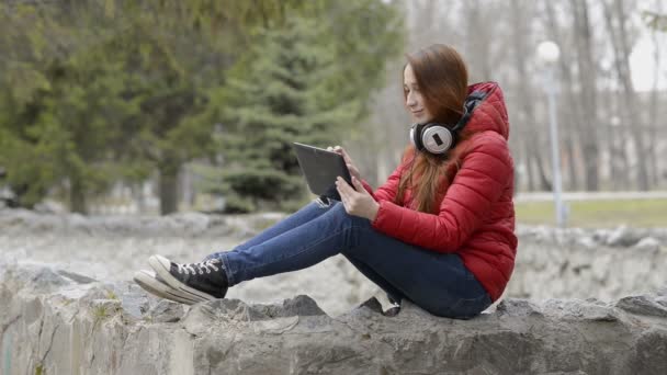 Hermosa chica descansando y divirtiéndose jugando en la tableta, sentado con auriculares en su cabeza en el parque de primavera de la ciudad. Tiene el pelo rojo y una chaqueta roja. Retrato. Vista lateral. 29,97 fps . — Vídeo de stock