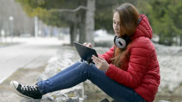 Uma jovem adolescente joga em um tablet, sentado em pedras perto de uma fonte vazia na cidade primavera Parque em uma jaqueta vermelha e com cabelo vermelho na cabeça. Retrato. Fecha. 29,97 fps . — Vídeo de Stock