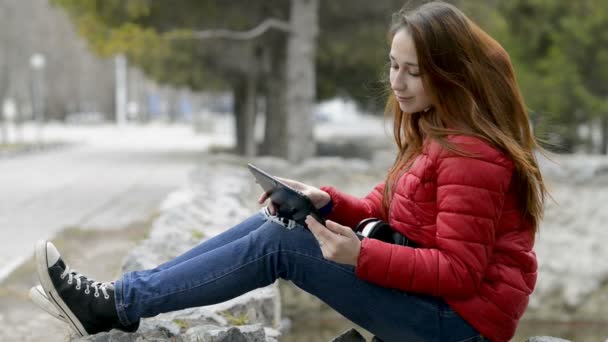 Ein trauriges junges Mädchen blickt auf den Bildschirm eines digitalen Tablets und richtet ihre Haare, sitzt in roter Jacke und mit roten Haaren auf den Steinen im Stadtpark. Nahaufnahme. 29,97 fps.