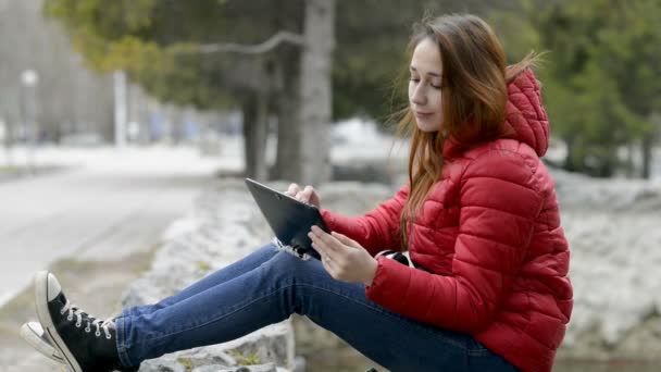 Słodka dziewczyna uśmiecha się i pisze wiadomość na ekranie cyfrowego tabletu, siedząc na skałach w parku miejskim wiosną w czerwonej kurtce i z czerwonymi włosami na głowie. Z bliska. Widok z boku. 29,97 fps. — Wideo stockowe