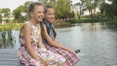İki neşeli küçük kız nehir kıyısında ahşap bir köprüye oturmak, gülmek ve gün batımı bir yaz akşamı bacaklarını salıncak. Portre. Kapat şunu. 4k. 25 fps.