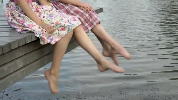 Los niños rápidamente balancean sus pies desnudos, sentados sobre el agua en un puente de madera en la orilla del río en una noche de verano. Primer plano. 4K. 25 fps . — Vídeos de Stock