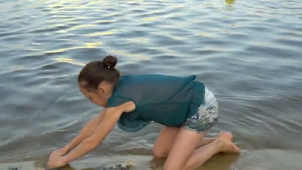 Malá holčička hraje na pláži při západu slunce a staví na pozadí klidné vody písečnou věž. Portrét. Zavři to. 4k. 25 fps.