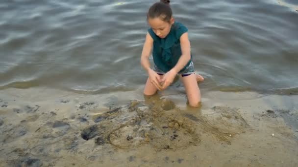 日落时, 一个小女孩在河岸上玩湿沙, 坐在膝盖上。她收集沙手在水边和建设。闭上你的嘴从顶部的视图。4k. 25 帧/秒. — 图库视频影像