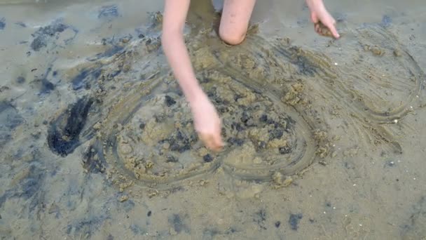 Una joven se dedica a la creatividad y dibuja un corazón en la arena húmeda en el borde del agua al atardecer, sentada sobre sus rodillas. La vista desde arriba. 4k. 25 fps . — Vídeos de Stock