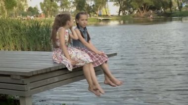 İki mutlu küçük kız gün batımında ahşap bir köprü üzerinde bir yaz akşamı şehir parkı nehir kıyısında oturuyor. Gülümsüyor, kameraya bakıp ellerini sallıyorlar. Close. 4k.