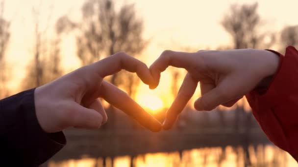 Un hombre y una niña enamorados sostienen sus manos en forma de corazón, símbolo del amor, sobre el fondo de una puesta de sol de color amarillo brillante sobre la naturaleza en la orilla del río. De cerca. 4K. 25 fps — Vídeo de stock