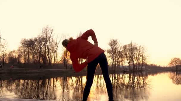Una joven se dedica a ejercicios de gimnasia al atardecer en las orillas del río en el parque de la ciudad. Vista trasera. 4K. 25 fps — Vídeos de Stock