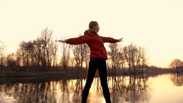 Młoda dziewczyna gra w sporcie w parku miejskim o zachodzie słońca. Wykonuje ćwiczenia gimnastyka i rozciąga się na brzegu rzeki w parku miejskim wiosną. Widok z tyłu. 4K. 25 kl./s — Wideo stockowe