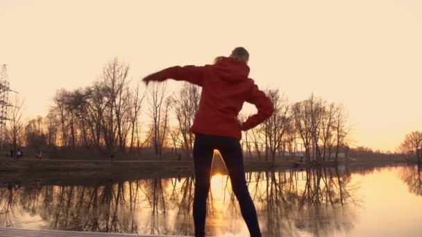 Athletic młoda dziewczyna zaangażowany w ćwiczenia gimnastyka i rozciąganie o zachodzie słońca na brzegu rzeki w parku wiosną miasta. Widok z tyłu. 4K. 25 kl./s — Wideo stockowe