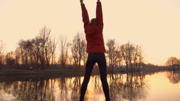 Chica joven haciendo deportes de pie en la orilla del río al atardecer en un parque de la ciudad con una chaqueta roja. Está iluminado por la luz del sol de color amarillo brillante. Vista trasera. 4K. 25 fps — Vídeos de Stock