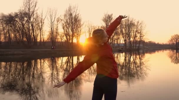 Primer plano de una joven que hace gimnasia de pie al atardecer en un parque de la ciudad. Se entrena en la orilla del río con una chaqueta roja e ilumina con luz solar de color amarillo brillante. Vista trasera. 4K. 25 fps — Vídeos de Stock