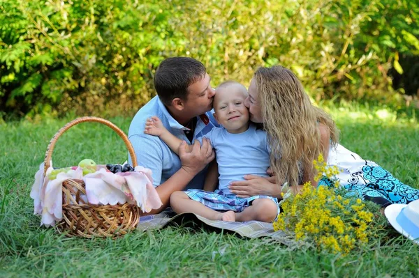 Um garotinho olha para a câmera e sorri, e mamãe e papai o beijam em ambos os lados, sentados na grama no parque no verão ao pôr-do-sol. Um piquenique familiar na natureza. Retrato — Fotografia de Stock