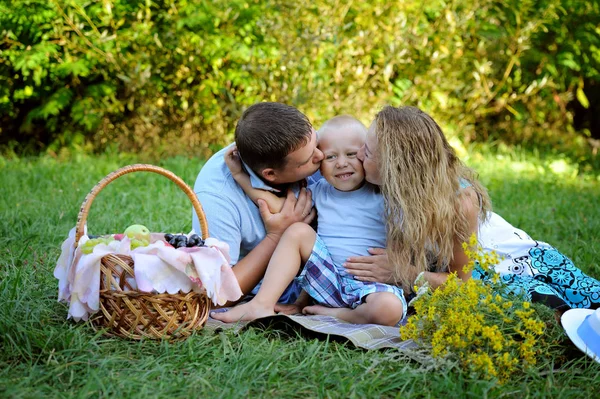 Mom and dad together kiss smiling little son sitting on the grass in the Park in the summer at sunset. Family picnic outdoors. Portrait. Horizontal orientation of the image — Stock Photo, Image