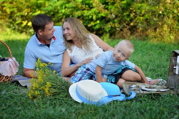 Family picnic in nature. Mom, dad and son relax sitting on the grass in the Park in the summer at sunset. A little boy looks at the camera and smiles. Portrait. Horizontal orientation of the image — Stock Photo, Image