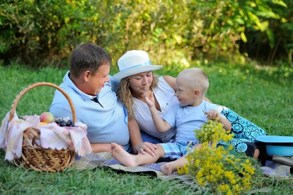 Petit garçon souriant nourrissant des raisins maman et papa assis sur l'herbe en été au coucher du soleil près d'un panier de fruits et un bouquet de fleurs sauvages jaunes. Pique-nique en famille à l'extérieur. Portrait — Photo