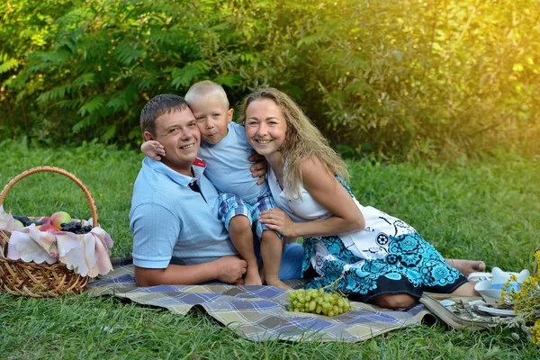 Feliz bebê abraços mãe e pai que estão sentados na grama no parque ao pôr do sol e sorrindo. Família piquenique ao ar livre. Retrato. Close-up. Orientação horizontal — Fotografia de Stock