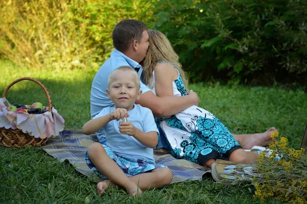 幸せな子供が草の上に座って微笑み、両親は日没時に公園で彼の後ろで抱きしめてキスをします。自然の中で家族の休暇。肖像 画。水平方向 — ストック写真
