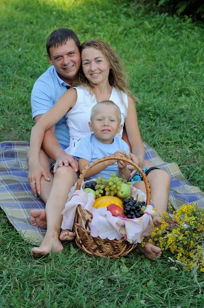 Pai, mãe e bebê se sentam um atrás do outro na grama no parque ao pôr do sol perto do cesto de frutas. Eles sorriem e olham para a câmera. Férias em família na natureza. Retrato. Vista vertical — Fotografia de Stock