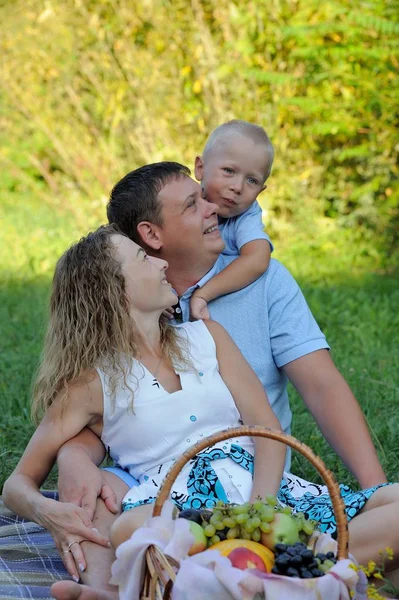 Cheerful little boy hugs his father and mother, who are sitting on the grass in the Park at sunset. They smile and look at the baby. Family vacation in nature. Portrait. Vertical view — Stock Photo, Image