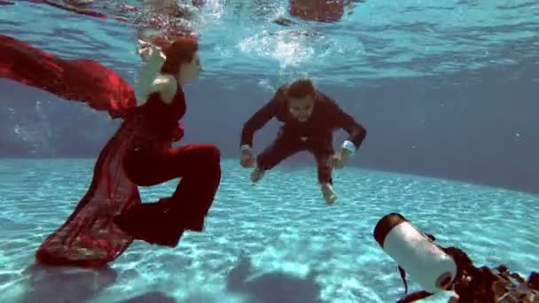 Подводный фотограф берет невесту и жениха в свадебных платьях под водой на дне бассейна на фоне света с водной поверхности. Медленное движение. Стрельба под водой. 4K — стоковое видео