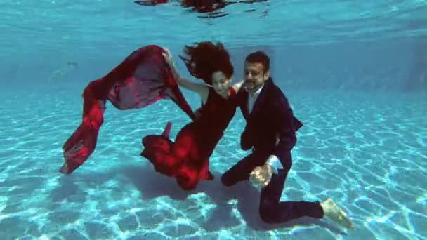 Novia y novio felices se sientan bajo el agua en el fondo de la piscina, sonriendo y mirando al fotógrafo que toma fotos de ellos. En cámara lenta. Fotografía submarina. 4K. 25 fps — Vídeo de stock