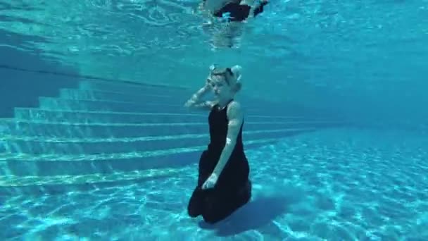 El fotógrafo submarino toma a una chica rubia con un vestido negro y cuernos blancos en su cabeza bajo el agua en el fondo de la piscina. Cosplay. En cámara lenta. Fotografía submarina. 4K. 25 fps — Vídeo de stock