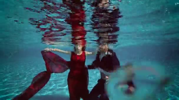 Uma noiva encantadora em um vestido vermelho e o noivo mergulhar sob a água para o fundo da piscina, sorriso e abraço. Movimento lento. Casamento tiroteio subaquático. 4K. 25 fps — Vídeo de Stock
