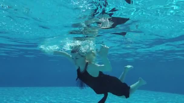 Молода блондинка плаває під водою біля поверхні в чорній сукні і прокидається у воді, руки витягнуті. Повільний рух. Вид знизу від басейну. 25 к/с — стокове відео