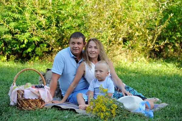 Papa, maman et le petit garçon sont assis sur l'herbe dans le parc un jour d'été, regardant loin et souriant. Pique-nique familial dans la nature. Portrait. Vue horizontale — Photo