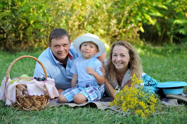 Un petit garçon dans un chapeau de femme sourit et s'assoit avec ses parents dans le parc sur l'herbe un jour d'été. Pique-nique familial dans la nature. Le concept de liberté de choix entre les sexes. Portrait. Vue horizontale — Photo