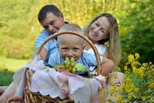 Un petit garçon regarde à travers un panier de fruits, et maman et papa s'assoient derrière lui sur l'herbe dans le parc, souriant et le regardant un jour d'été. Pique-nique familial dans la nature. Portrait. Vue horizontale — Photo