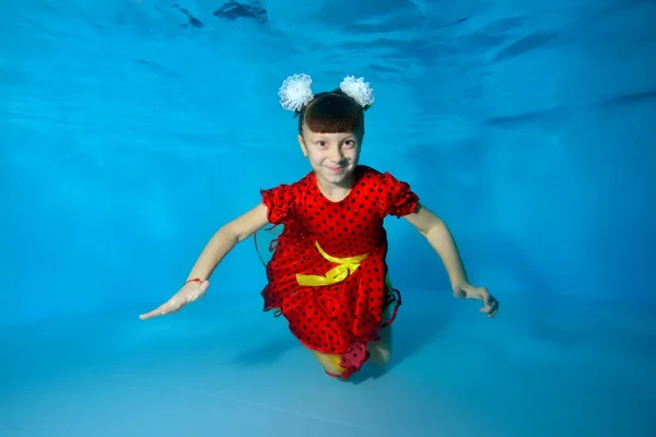 Una niña encantadora nada bajo el agua con los ojos abiertos, sonríe y posa para la cámara con los brazos extendidos con un vestido rojo sobre un fondo azul. Retrato. Vista horizontal —  Fotos de Stock