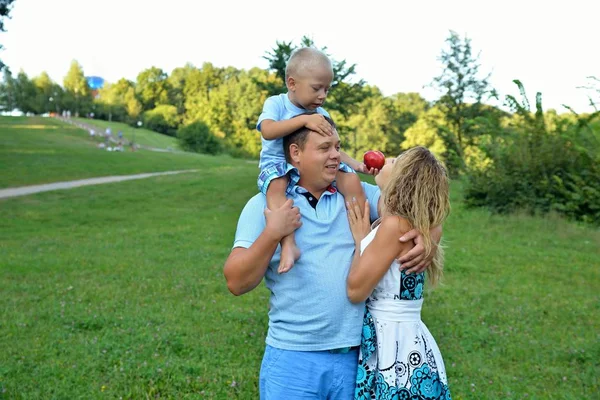 Un petit garçon traite sa mère avec une pomme rouge, assis sur le cou de son père dans le parc par une journée d'été ensoleillée sur un fond vert. Activités familiales de plein air. Le concept de bonheur familial — Photo