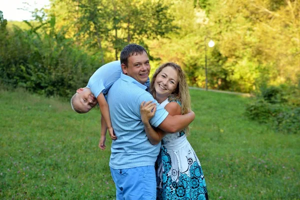 Famille heureuse : papa, maman et petit fils marchent et jouent dans le parc à l'extérieur. Papa tient le bébé sur son épaule, ils sourient tous ensemble et regardent la caméra. Le concept de bonheur familial — Photo