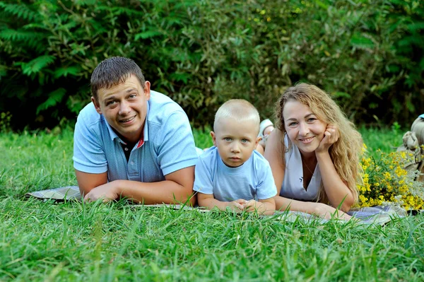Portrait de père, mère et enfant souriants. Ils s'allongent sur le ventre dans le parc sur l'herbe au coucher du soleil et me regardent. Vacances en famille dans la nature. Vue horizontale — Photo