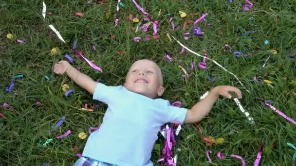 Leende blond liten pojke som ligger på det gröna gräset, tittar upp och vinka vid solnedgången av en sommardag i parken. Runt barnet spridda glitter från fyrverkerierna. Koncept. 4K. 25 fps — Stockvideo