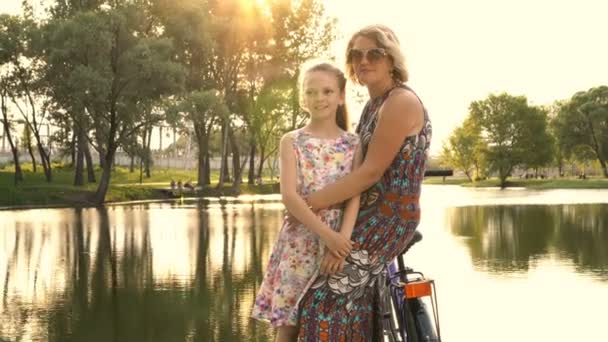 Счастливая мама и маленькая дочь в красивых платьях обнимаются на закате летнего дня на фоне реки в парке. Они стоят и опираются на фиолетовый велосипед у воды. Концепция. 4К. 25 кадров в секунду — стоковое видео