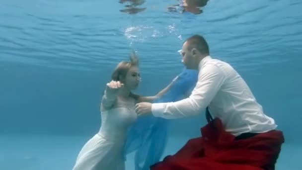 The bride in white dress and the groom swim and play underwater in the pool with red and blue fabric. Wedding underwater. Portrait. Zoom. 4K. 29.97 fps — Stock Video