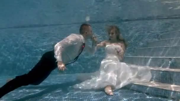 Novia feliz y novio nadan y juegan en la parte inferior de la piscina en trajes de novia blancos en un día soleado. El tipo besa a las chicas bajo el agua. Boda bajo el agua. Retrato. 4K. 29,97 fps — Vídeos de Stock