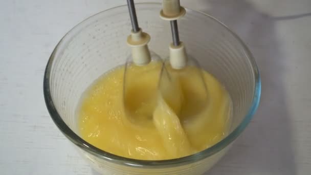 Close-up, um misturador elétrico chicoteia ovos em uma tigela de vidro em uma mesa branca para cozinhar. A vista de cima. 4K. 25 fps — Vídeo de Stock