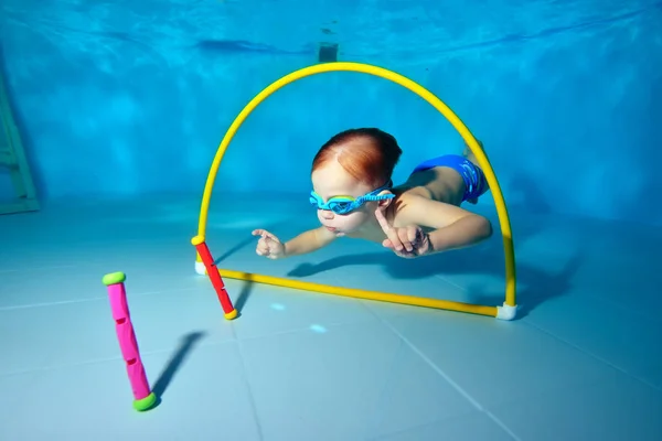 Un niño pequeño bucea y nada bajo el agua en la piscina para juguetes a través de un aro amarillo. Retrato. Fotografía submarina. Orientación horizontal —  Fotos de Stock