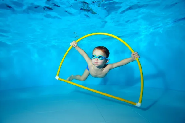 Un niño pequeño se dedica a nadar bajo el agua en la piscina cerca del fondo, posando con un aro amarillo en las manos y mirando hacia arriba. Retrato. Fotografía submarina. Orientación horizontal — Foto de Stock