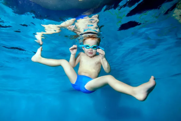 Un niño nada y juega bajo el agua en la piscina con las piernas extendidas hacia los lados y mira a la cámara. Retrato. Fotografía submarina. Orientación horizontal de la imagen — Foto de Stock