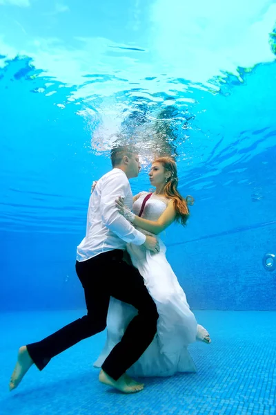 Lycklig brud och brudgum stå i bröllopsklänningar under vattnet längst ner i poolen, kramas och titta på varandra. Porträtt. Koncept. Undervattensfotografering. Vertikal vy — Stockfoto