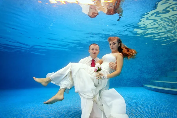 Hält der Bräutigam die Braut am Grund des Pools unter Wasser in den Armen. die Braut hält Blumen in der Hand. Porträt. Konzept. Unterwasserfotografie. horizontale Ansicht — Stockfoto